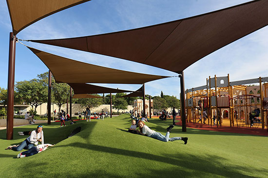 People are relaxing on a hilly grass area next to a large playground. They are shaded with large tension sail shade installations above the grassy area.